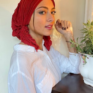 May include: A red ruffled turban head wrap made of soft fabric. The turban is worn by a person with a white button-down shirt.
