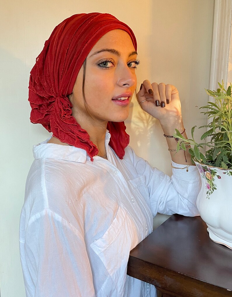 May include: A red ruffled turban head wrap made of soft fabric. The turban is worn by a person with a white button-down shirt.