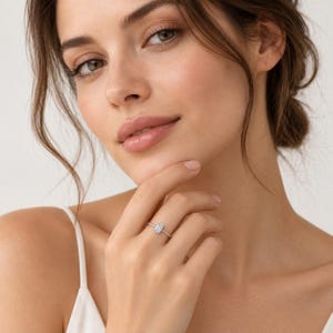 Puede incluir: Un anillo de tono plateado con una sola piedra preciosa transparente de corte esmeralda montada sobre una banda de metal fina y pulida. El anillo se lleva en un dedo, destacando las facetas de la piedra rectangular.
