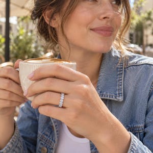 Puede incluir: Un anillo de tipo alianza en tono plateado con una hilera continua de piedras transparentes de talla ovalada engastadas en una banda de metal pulido, que se lleva en el dedo mientras se sujeta una taza de cerámica.