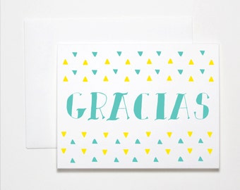 May include: A white card with a teal and yellow triangle pattern on the top and bottom. The word "GRACIAS" is written in teal in the center of the card.