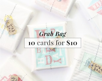 May include: A collection of 10 greeting cards bundled together and tied with twine. The cards are wrapped in white tissue paper and have a variety of designs. The text "Grab Bag 10 cards for $10" is printed on a white rectangle in the center of the image.