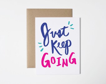 May include: A white card with the text "Just Keep Going" in blue and pink lettering. The text is written in a hand-lettered style. The card has a brown envelope.