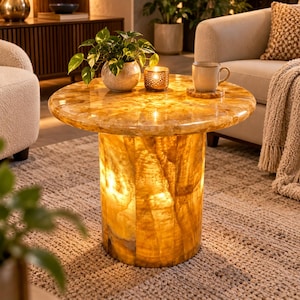 May include: A cylindrical side table crafted from translucent golden-yellow stone with a polished finish. The table features an illuminated base that emits a warm glow, topped with a potted green plant, a metallic candle holder, and a ceramic mug.