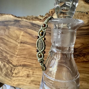 May include: A macrame bracelet featuring light green cord, brown tiger eye beads, and a central metallic leaf charm. The bracelet is draped over the neck of a clear glass decanter with a faceted stopper, showcasing intricate knotting.