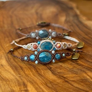 May include: Three macrame bracelets featuring central faceted blue stones, smaller accent beads, and knotted cord bands. The bracelets include metallic heart-shaped charms and adjustable closures, showcasing intricate woven patterns in brown and cream tones.