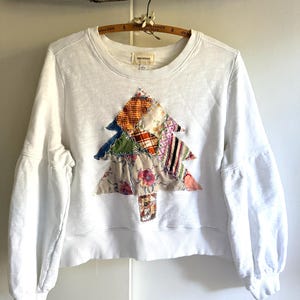 May include: A white long-sleeve sweatshirt featuring a central patchwork Christmas tree design. The tree is constructed from various fabric scraps in plaid, floral, and striped patterns with raw, frayed edges. The garment has a crew neckline.