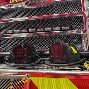 May include: Two black fire helmets with red leather front shields featuring the number 91 and the names DOWELL and MAYO, positioned in front of a red fire truck with a chrome grille and the word Pierce visible on the front.