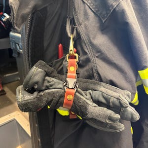 May include: A brown leather glove strap with a black plastic side-release buckle and a brass swivel snap hook. The strap is attached to a black jacket with yellow reflective trim, holding a pair of black textured work gloves in place.