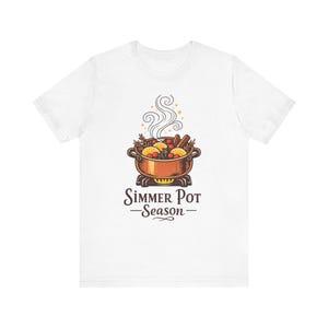 Può includere: Una t-shirt bianca a maniche corte con la grafica di una pentola color rame su un fornello. La pentola contiene fette d'arancia, mirtilli rossi e bastoncini di cannella con vapore che sale. Il testo recita Simmer Pot Season.