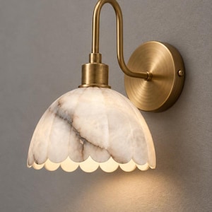 May include: A wall-mounted light fixture featuring a curved brass arm and a circular brass wall plate. The lamp shade is crafted from translucent white stone with natural grey veining, shaped into a scalloped dome that emits a soft glow.