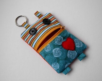 Op de afbeelding: Een klein, blauw en oranje stoffen tasje met een ritssluiting en een hartvormige patch. Het tasje heeft een sleutelhanger en twee stoffen lussen aan de zijkanten.
