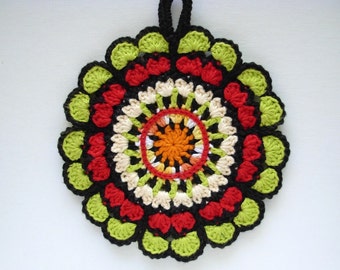 Op de afbeelding: Een gehaakte pot onderzetter met een cirkelvormig ontwerp met zwart, rood, groen, wit en oranje garen.