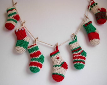 Op de afbeelding: Een slinger van kleine, gehaakte kerstsokken in rood, groen en crème. De sokken zijn geregen aan een touw en vastgezet met kleine houten knijpers. Een feestelijke decoratie voor de feestdagen.