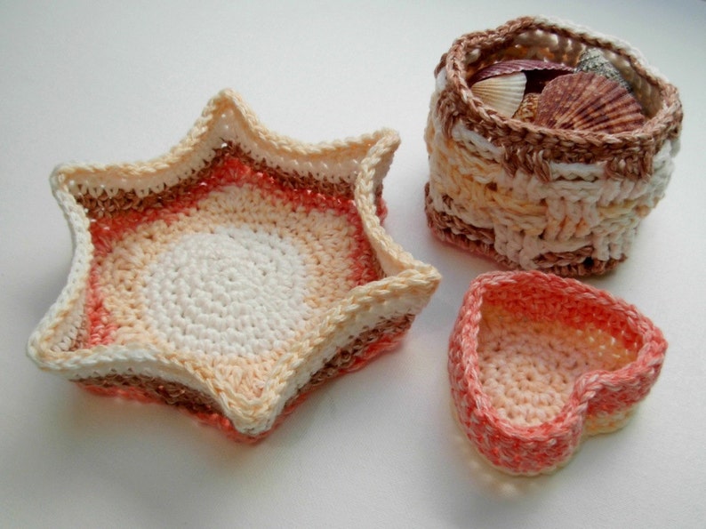 Op de afbeelding: Drie gehaakte manden in verschillende vormen: een ster, een hart en een ronde mand. De ster- en hartmanden zijn in tinten roze en wit, terwijl de ronde mand in tinten bruin, beige en wit is. De ronde mand is gevuld met schelpen.