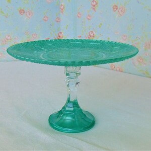 May include: A teal green glass cake stand with a scalloped edge and a clear glass pedestal.
