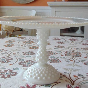 May include: A white hobnail cake stand with a round, beaded edge plate. The stand has a textured, bulbous stem and a wide, round base. The cake stand is on a floral patterned tablecloth.