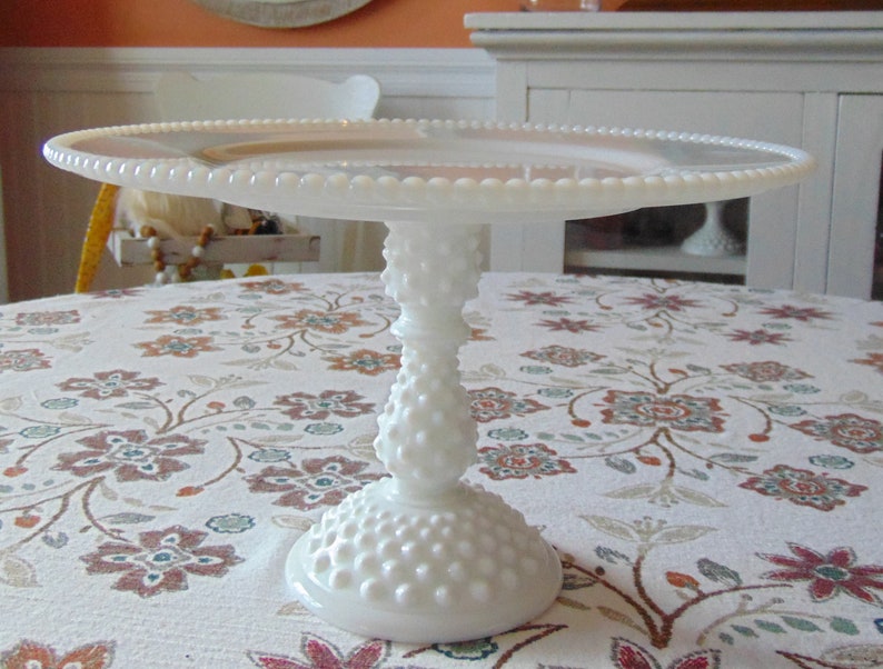 May include: A white hobnail cake stand with a round, beaded edge plate. The stand has a textured, bulbous stem and a wide, round base. The cake stand is on a floral patterned tablecloth.