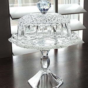 May include: A clear glass cake stand with a diamond-shaped knob on the lid. The stand has a pedestal base and a decorative, scalloped edge. The glass has a cut-crystal design, reflecting light.