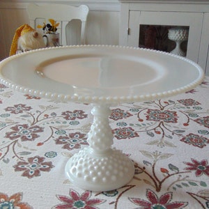 May include: A white milk glass cake stand with a beaded edge and a textured stem. The stand is round and elevated, perfect for displaying baked goods. The stand is on a floral tablecloth.