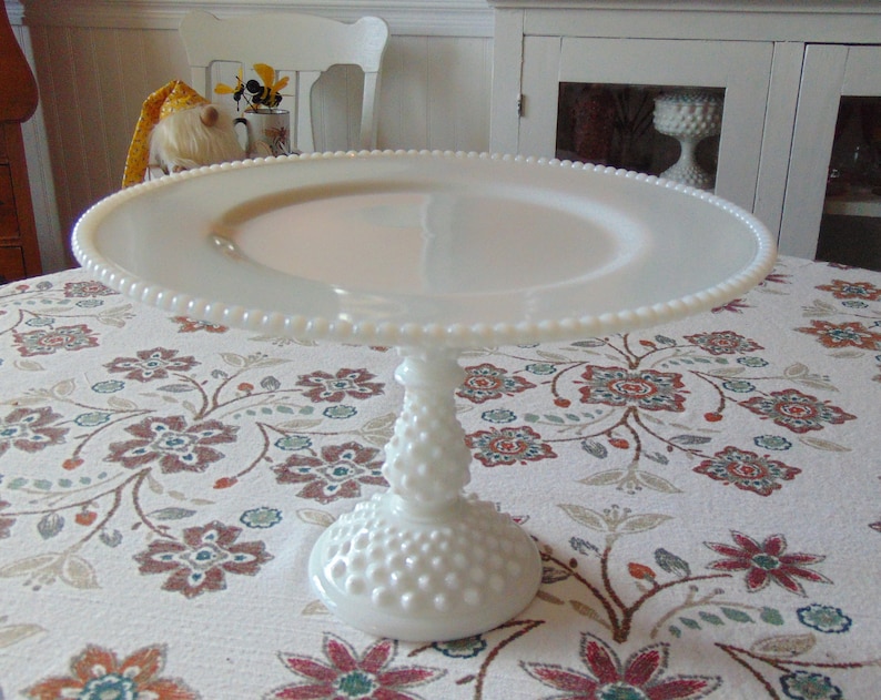 May include: A white milk glass cake stand with a beaded edge and a textured stem. The stand is round and elevated, perfect for displaying baked goods. The stand is on a floral tablecloth.