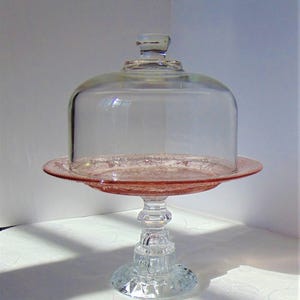 Puede incluir: Un soporte para pasteles de cristal rosa vintage con una c&uacute;pula de cristal transparente. El soporte tiene una base y un tallo decorativos, y el plato tiene un borde detallado. La c&uacute;pula tiene un peque&ntilde;o asa en la parte superior.