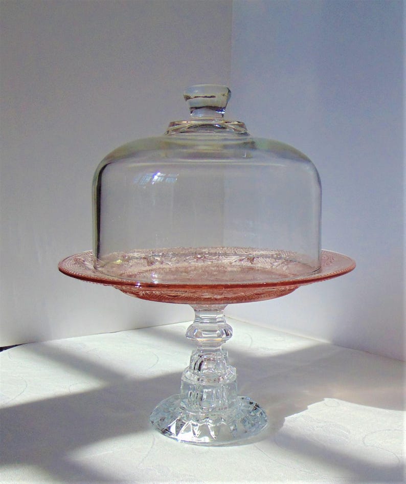 Puede incluir: Un soporte para pasteles de cristal rosa vintage con una c&uacute;pula de cristal transparente. El soporte tiene una base y un tallo decorativos, y el plato tiene un borde detallado. La c&uacute;pula tiene un peque&ntilde;o asa en la parte superior.