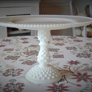 May include: A white, hobnail-style cake stand with a round top and a pedestal base. The stand is made of glass or ceramic and features a beaded edge. The stand is on a floral patterned tablecloth.