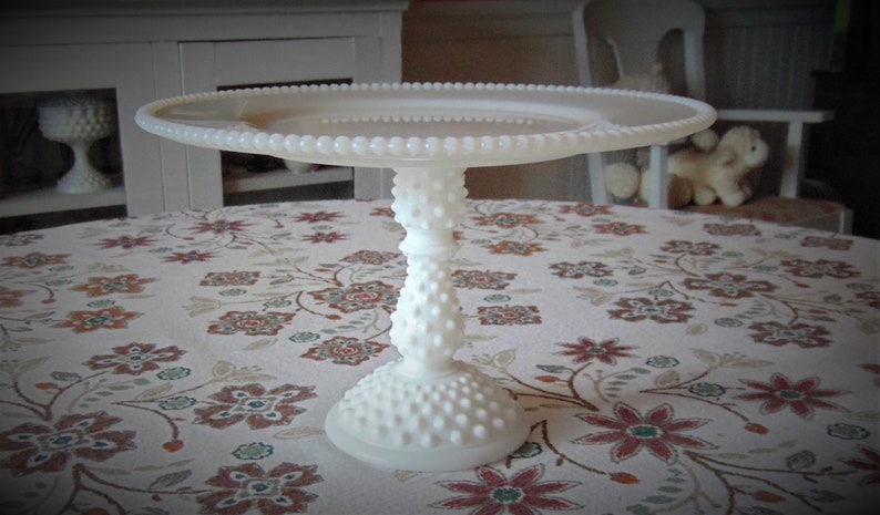 May include: A white, hobnail-style cake stand with a round top and a pedestal base. The stand is made of glass or ceramic and features a beaded edge. The stand is on a floral patterned tablecloth.