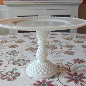 May include: A white, vintage-style cake stand with a round, beaded edge and a textured, hobnail-like base and stem. The cake stand is displayed on a floral patterned tablecloth, showcasing its elegant design. The stand measures approximately 12 inches in diameter.