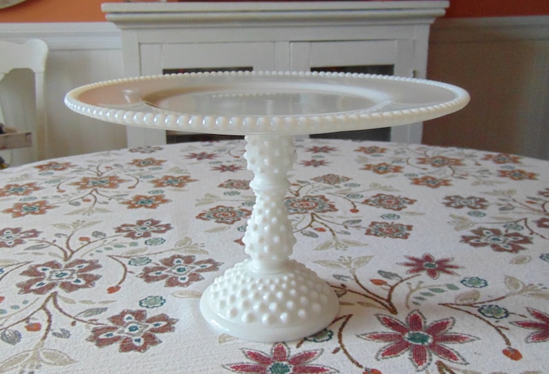 May include: A white, vintage-style cake stand with a round, beaded edge and a textured, hobnail-like base and stem. The cake stand is displayed on a floral patterned tablecloth, showcasing its elegant design. The stand measures approximately 12 inches in diameter.
