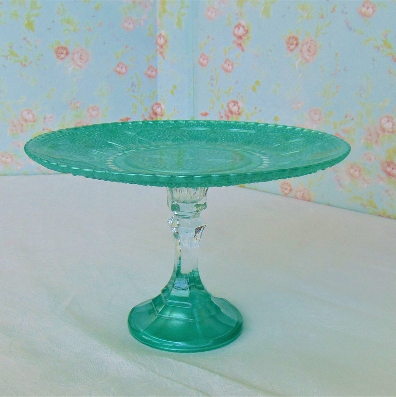 May include: A teal green glass cake stand with a scalloped edge and a clear glass pedestal.