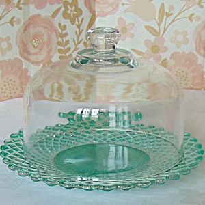 May include: A clear glass cake stand with a domed lid. The base and lid are a light teal color. The base has a scalloped edge and a diamond pattern. The lid has a clear knob on top.