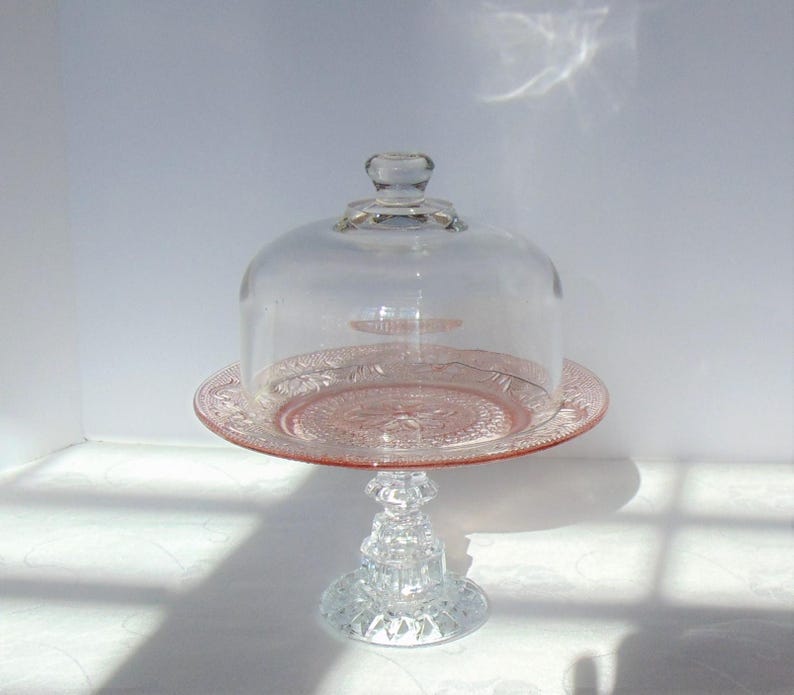 Puede incluir: Un soporte para pasteles de cristal rosa vintage con una c&uacute;pula de cristal transparente. El soporte tiene una base detallada y ornamentada y un patr&oacute;n decorativo en relieve en el plato. La c&uacute;pula tiene un peque&ntilde;o asa en la parte superior.