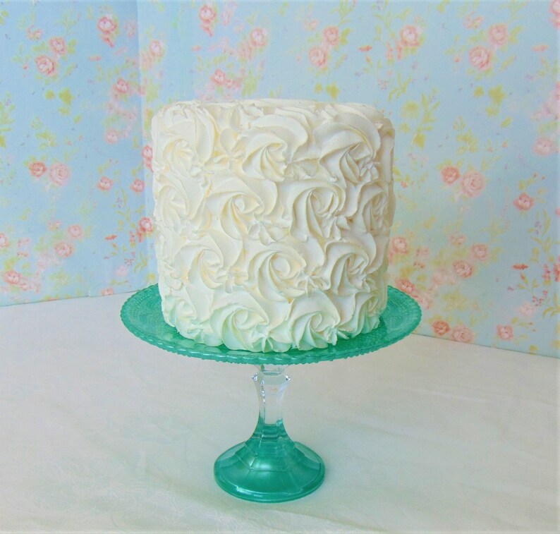 May include: A white cake with a rosette frosting design sits on a teal cake stand. The cake is on a white surface with a floral patterned background.