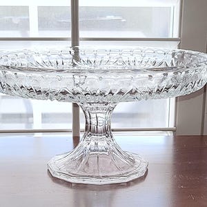 May include: A clear, pedestal-style glass serving dish with a decorative, textured pattern. The dish has a wide, shallow bowl and a sturdy base. The glass is transparent, allowing light to pass through, and is suitable for serving desserts or displaying decorative items.