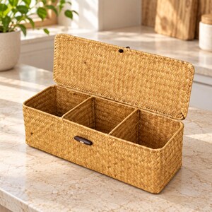 May include: A rectangular woven storage box with a hinged lid and three internal compartments. The box is crafted from light brown natural fibers and features a small, dark brown wooden toggle closure on the front center.