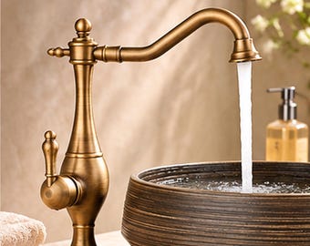 May include: A tall, antique brass-finished faucet with a curved spout and a single side lever handle. Water flows steadily from the spout into a dark, textured, bowl-shaped basin. The faucet features a decorative finial at the top.