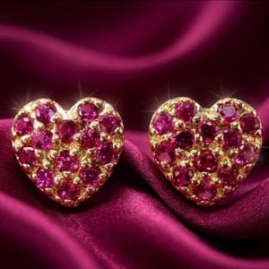 May include: A pair of heart-shaped stud earrings crafted from gold-toned metal, each featuring a cluster of small, round, vibrant pink stones set closely together to form a dense, sparkling surface across the entire heart shape.