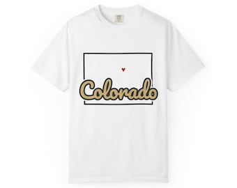 May include: A white short-sleeve t-shirt featuring a black rectangular outline. Inside the box is the word Colorado written in a tan cursive font with a black outline, positioned below a small red heart centered at the top.