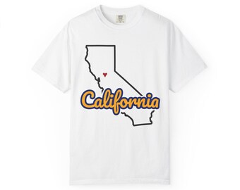 May include: A white short-sleeve t-shirt featuring a black outline of the state of California with a small red heart placed in the central region. The word California is printed in a yellow cursive font with a dark blue outline below.