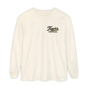 May include: An off-white long-sleeve crewneck shirt featuring a small black graphic on the upper left chest. The graphic displays the word Tigers in a cursive script font above the word FOOTBALL in a bold, block capital letter font.