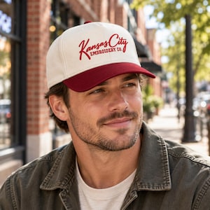 May include: A cream-colored baseball cap with a dark red brim. The front of the cap features the text Kansas City EMBROIDERY CO. in a dark red script and block font. The cap has a structured crown and a button on the top center.