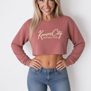 May include: A dusty rose long-sleeve crop top sweatshirt featuring a crew neckline and ribbed cuffs. The center of the chest displays the text Kansas City FOOTBALL CLUB in a light cream-colored script and block font combination.