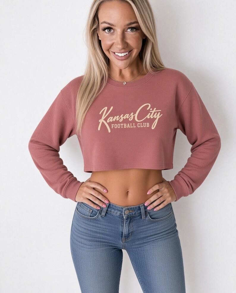 May include: A dusty rose long-sleeve crop top sweatshirt featuring a crew neckline and ribbed cuffs. The center of the chest displays the text Kansas City FOOTBALL CLUB in a light cream-colored script and block font combination.