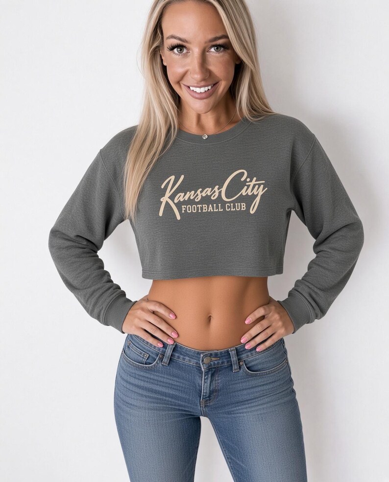 May include: A dark olive green long-sleeve crop top sweatshirt featuring a crew neckline and ribbed cuffs. The front displays the text Kansas City Football Club in a light beige script and block font. The garment has a textured fabric finish.