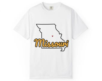 May include: A white short-sleeve t-shirt featuring a black outline of the state of Missouri. A small red heart is positioned in the centre of the state shape. The word Missouri is printed in a yellow cursive font with a black outline below.