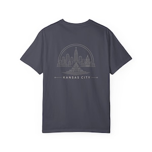Puede incluir: Una camiseta gris oscuro de manga corta con un gráfico de líneas blancas en la espalda. El diseño muestra el horizonte de Kansas City dentro de un semicírculo, con el texto KANSAS CITY impreso debajo en una fuente sencilla.