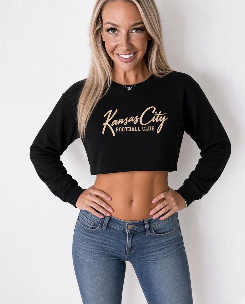 May include: A black long-sleeve cropped sweatshirt featuring the text Kansas City FOOTBALL CLUB in a tan script and block font across the chest. The garment has a crew neckline and ribbed cuffs, providing a fitted look for the upper body.