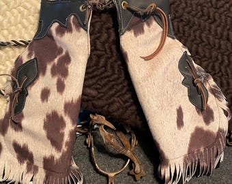 May include: A pair of chaps featuring a brown and white cowhide pattern with black leather trim, brass eyelets, and leather lacing. The chaps include decorative black leather accents on the sides and long fringe along the bottom edges, accompanied by a metal spur.